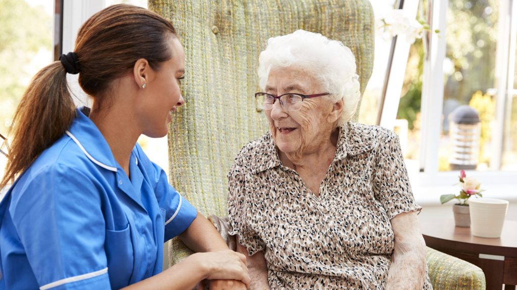 Senior Woman Sitting In Chair And Talking With Nurse In Retirement Home